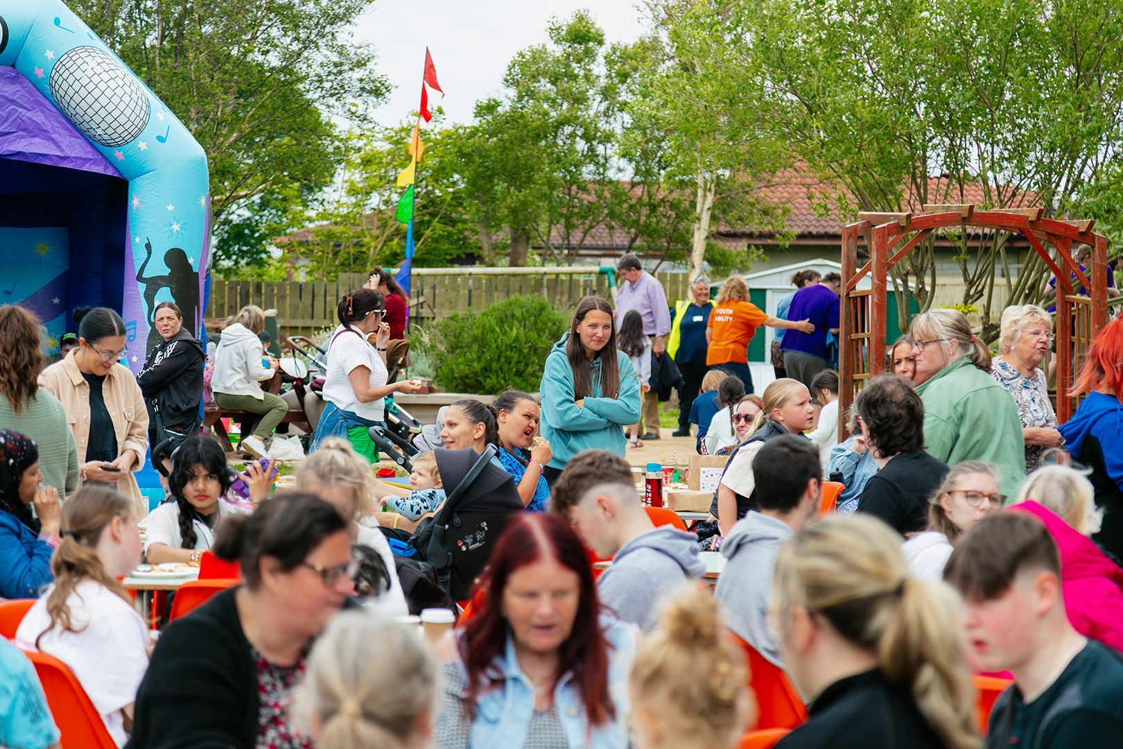 A crowd of people gathered for the Love Barrow Libraries Big Lunch 2024, sitting at tables enjoying a Big Lunch outside. There is a blue and purple bouncy castle in the background, along with trees. A young woman in a blue top is looking at the camera and smiling.