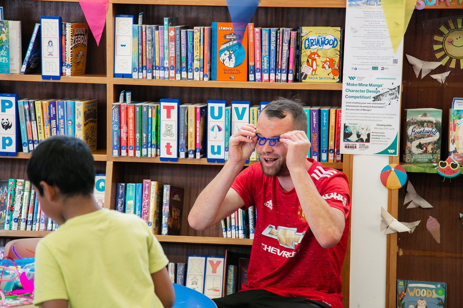 A man at the Love Barrow Libraries Big Lunch is joking around and making glasses out of some blue pipe cleaners whilst sitting in the library in front of some books. He looks like he is laughing and having a good time.