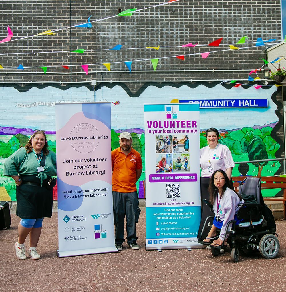 2 women a man and a lady in a wheelchair are posing with a Love Barrow Libraries banner and volunteering banner outside of Walney Library in Barrow