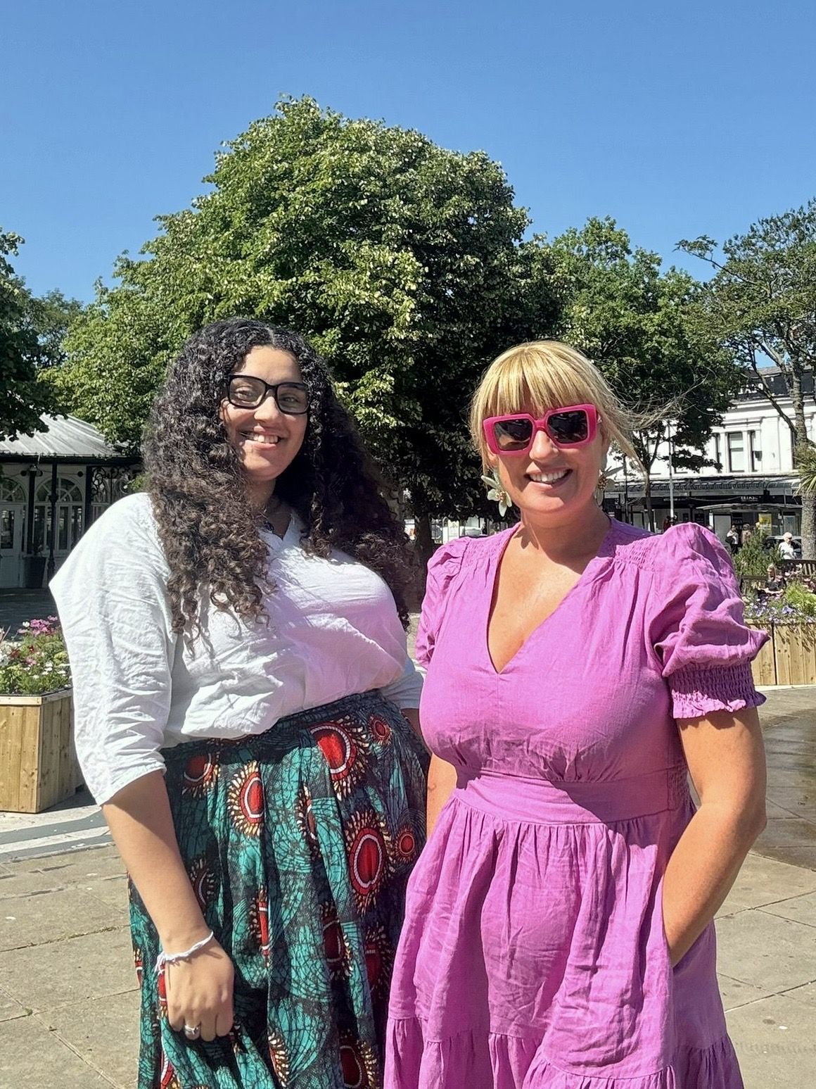 Two women dressed in bright and flowing outfits have their arms round each other and are smiling - the sky is blue in the background