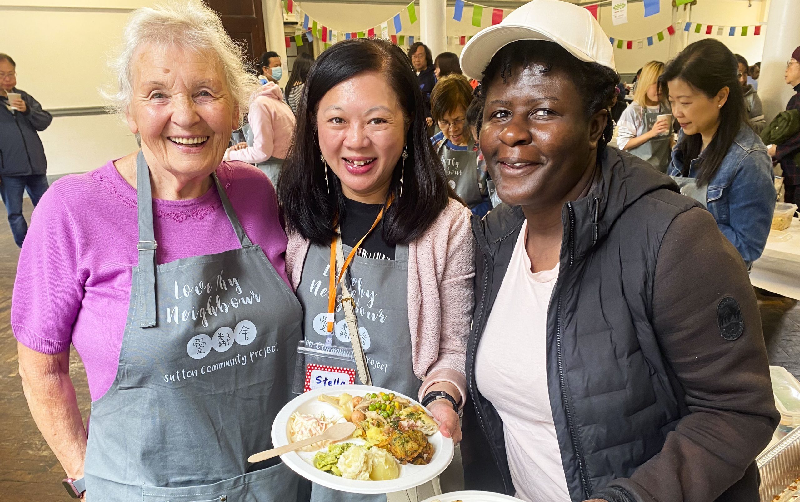 Three women from the Sutton Community Project are sharing plates of homemade food together, looking happy. Two of them are wearing cooking aprons.