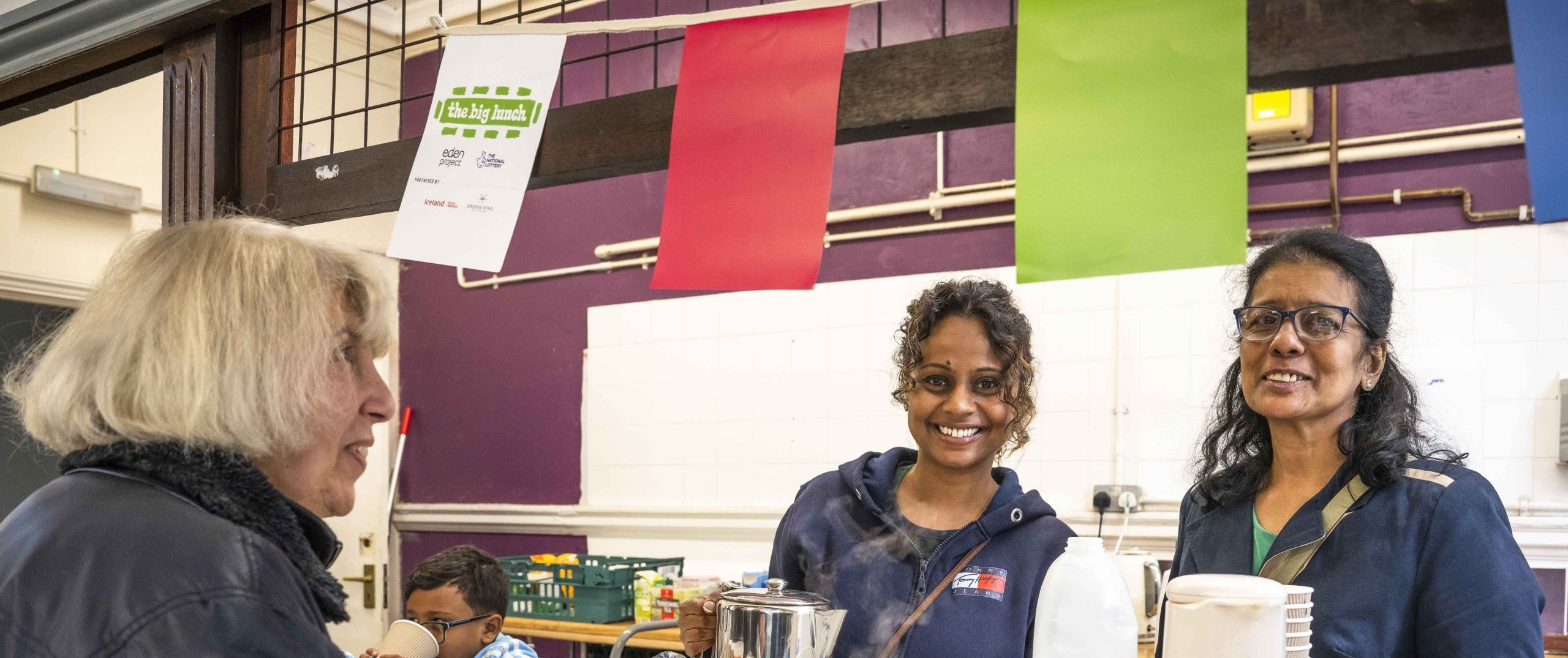 Two smiling women are serving another woman and drink and food at the Sutton Community Project Big Lunch. There is colourful bunting hanging.