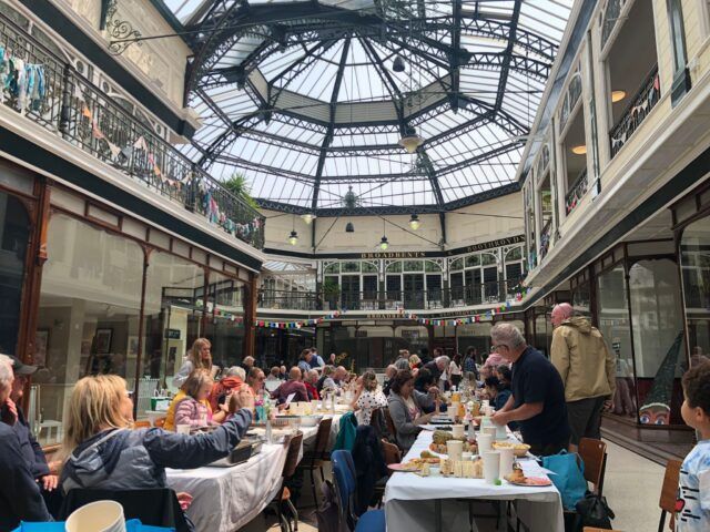 A Big Lunch spread - two tables of people chatting to each other with delicious food in front of them. They're in a Victorian shopping arcade with glass dome ceiling.