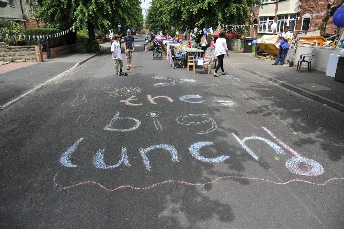 The Big Lunch is written in colourful chalk on the road. In the background children and adults are playing and sitting at trestle tables in the street.