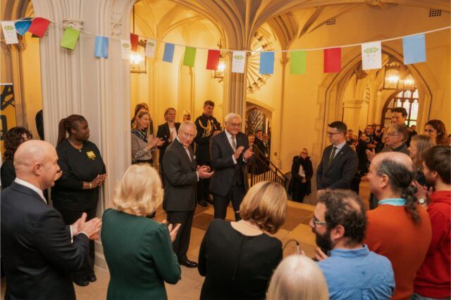 HM The King and The Federal President of Germany standing together, with people surrounding. There is bunting overhead.