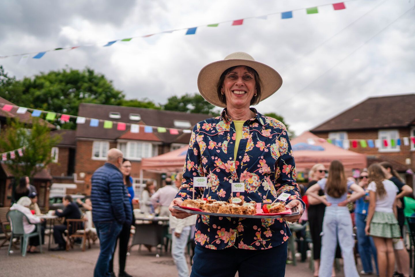 A woman stands beaming with a tray of cakes, with a street party taking place in the background