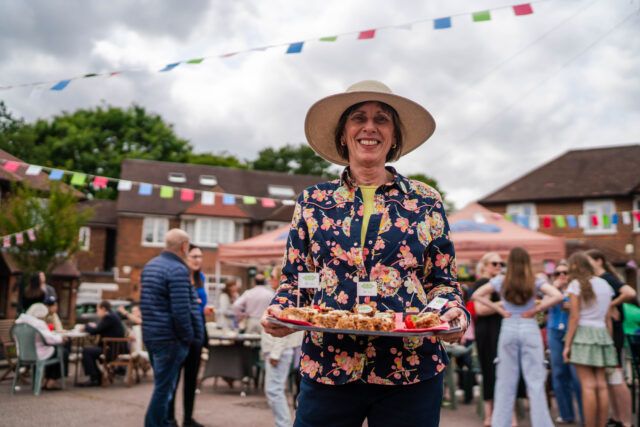 A woman stands beaming with a tray of cakes, with a street party taking place in the background