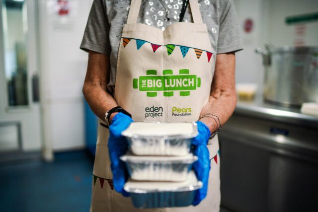 A woman wearing an apron branded The Big Lunch carries three takeaway containers