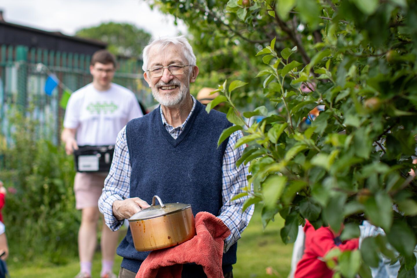 Man carrying saucepan through allotment. He's smiling and more people are visible behind carrying things