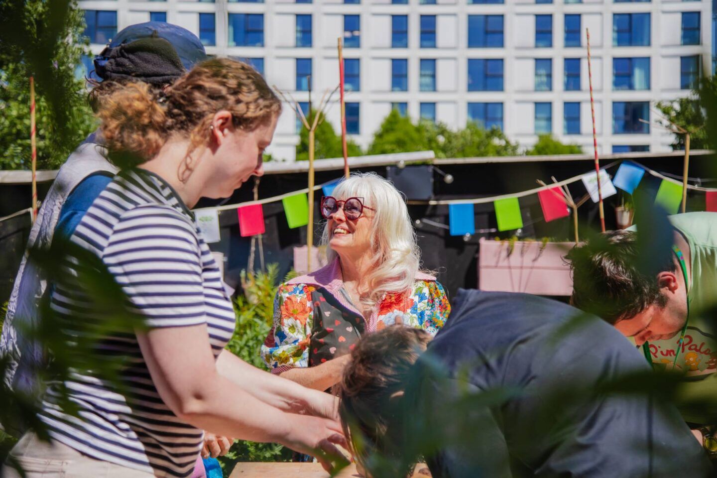 A woman smiles up at another person next to her. She's standing in a group all doing an activity in their hands we can't quite see. It's sunny and warm with colourful bunting in the background.