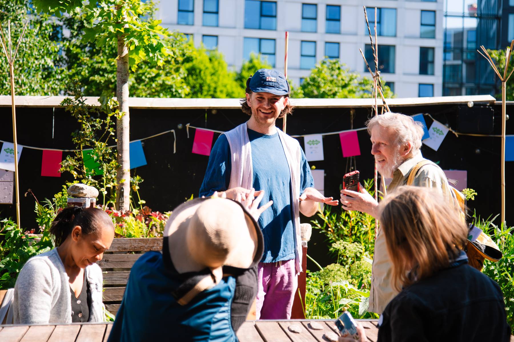 A man smiles in front of a group of people at a sunny Big Lunch. They're in a greenspace in an urban setting.