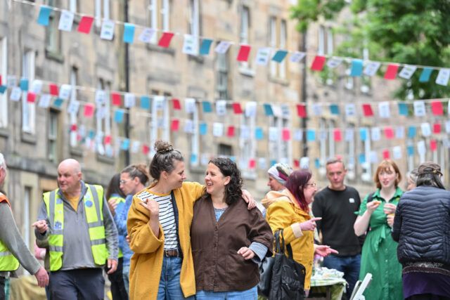 Two women stand with their arms around each other in front of a group of chatting people at a street party. Bunting is strung up across the street above their heads.