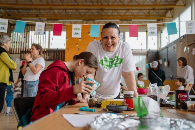 A woman wearing a Big Lunch t-shirt smiles at the camera, standing next to a young girl doing a craft activity.