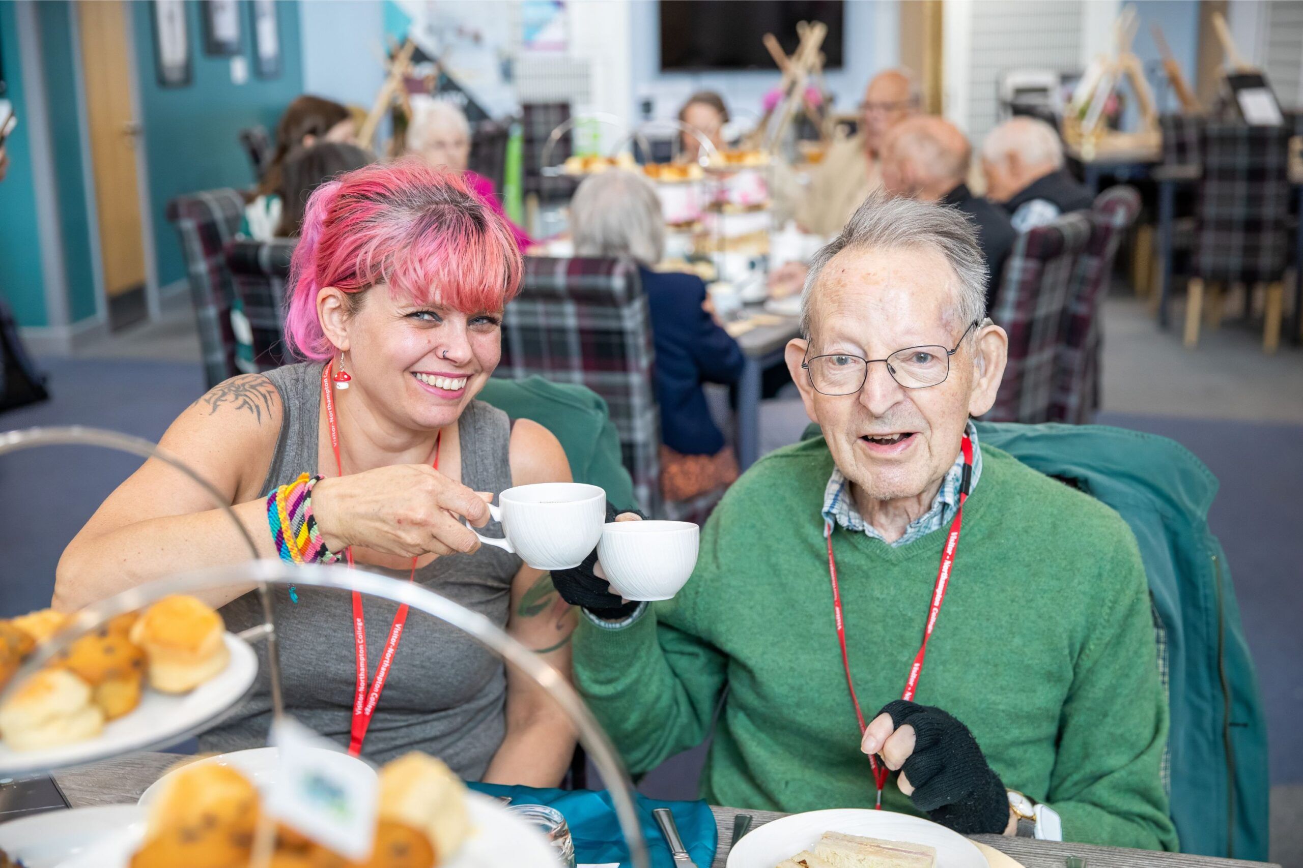 A young woman with pink hair and an elderly man make a cheers motion with a cup of tea